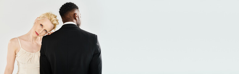 A beautiful blonde bride in a wedding dress and an African American groom stand side by side in a studio against a grey background.