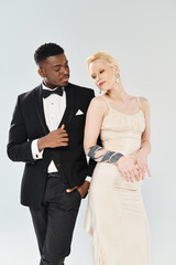 An African American groom in a tuxedo and a beautiful blonde bride in a wedding dress standing together in a studio on a grey background.