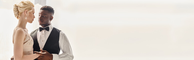 A beautiful blonde bride in a wedding dress and an African American groom stand next to each other in a studio against a grey background.
