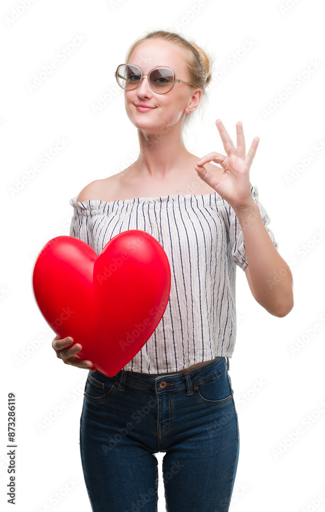 Poster blonde teenager woman holding red heart doing ok sign with fingers, excellent symbol