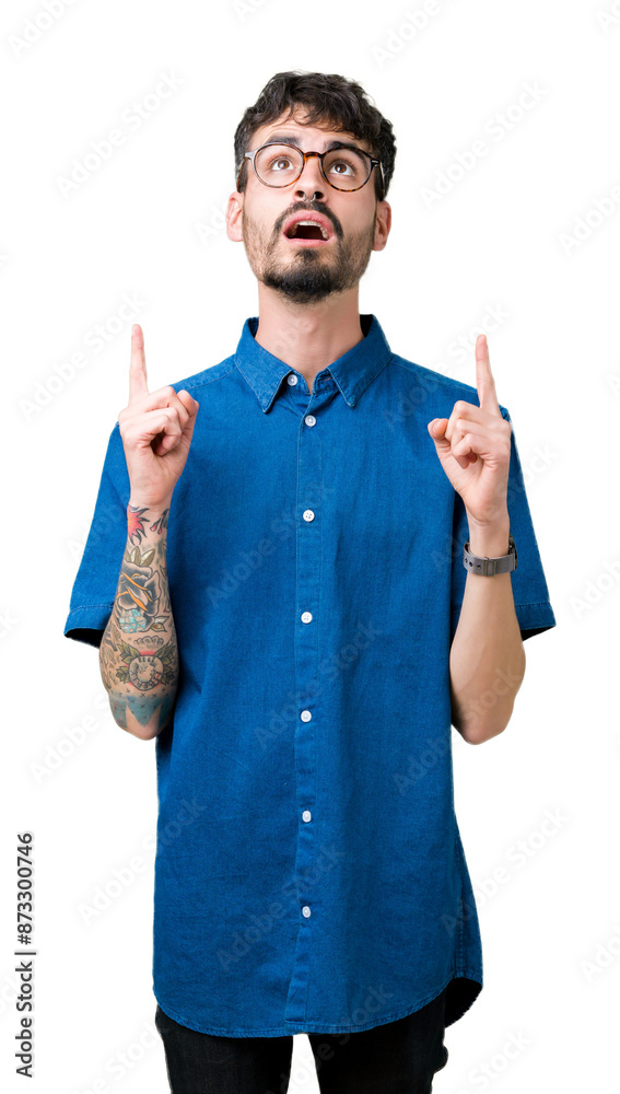 Sticker Young handsome man wearing glasses over isolated background amazed and surprised looking up and pointing with fingers and raised arms.