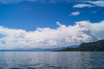 A view of Lake Garda. Interesting cloud formations are reflected on the lake, with the surrounding hills and mountains on the horizon. 