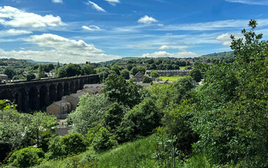 A serene rural landscape features lush greenery, a stone viaduct, and quaint buildings under a partly cloudy sky, with an extensive view of rolling hills and forested areas in, Milnsbridge, UK