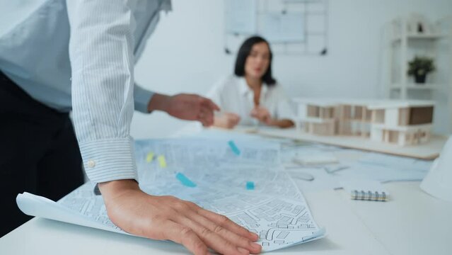 Skilled caucasian interior designer working with house model while discussing with asian architect engineer about house plan on meeting table with blueprint and house model scatter around. Immaculate.