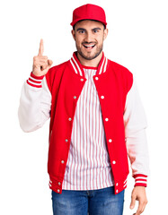 Young handsome man wearing baseball jacket and cap pointing finger up with successful idea. exited and happy. number one.