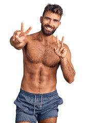 Fototapeta premium Young hispanic man wearing swimwear shirtless smiling looking to the camera showing fingers doing victory sign. number two.