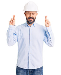Young handsome man wearing architect hardhat gesturing finger crossed smiling with hope and eyes closed. luck and superstitious concept.