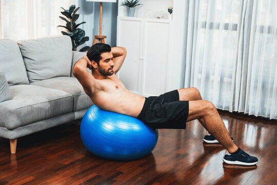 Athletic and sporty man doing situp on fitness ball during home body workout exercise session for fit physique and healthy sport lifestyle at home. Gaiety home exercise workout training concept.