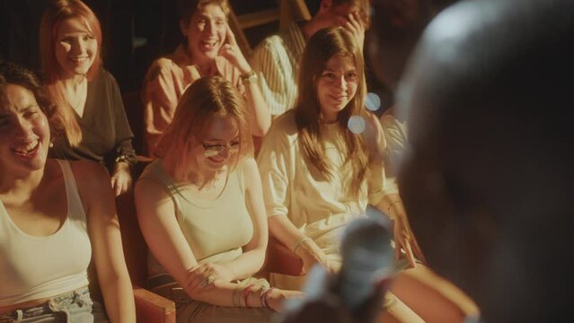 African American comedian telling jokes in microphone during stand-up performance on stage, people sitting in audience applauding and laughing. Close-up shot, rack focus