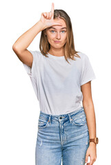 Beautiful young blonde woman wearing casual white t shirt making fun of people with fingers on forehead doing loser gesture mocking and insulting.