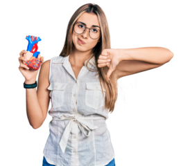 Hispanic young woman holding heart organ with angry face, negative sign showing dislike with thumbs down, rejection concept