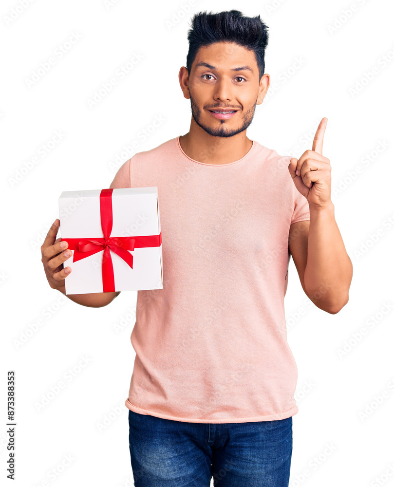 Wall mural Handsome latin american young man holding gift surprised with an idea or question pointing finger with happy face, number one