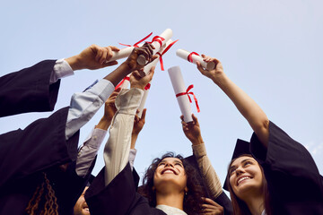 Happy graduate students standing in a circle in black robes and raising their diplomas up to the...