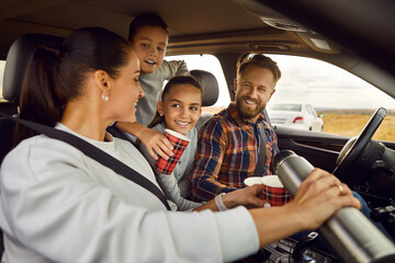 Joyful woman pouring hot drink from thermos into cups for her family during car trip. Parents and...