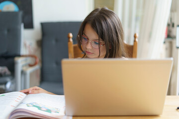 A little teenage girl in glasses does her homework on a laptop while sitting at the table. Homeschooling concept