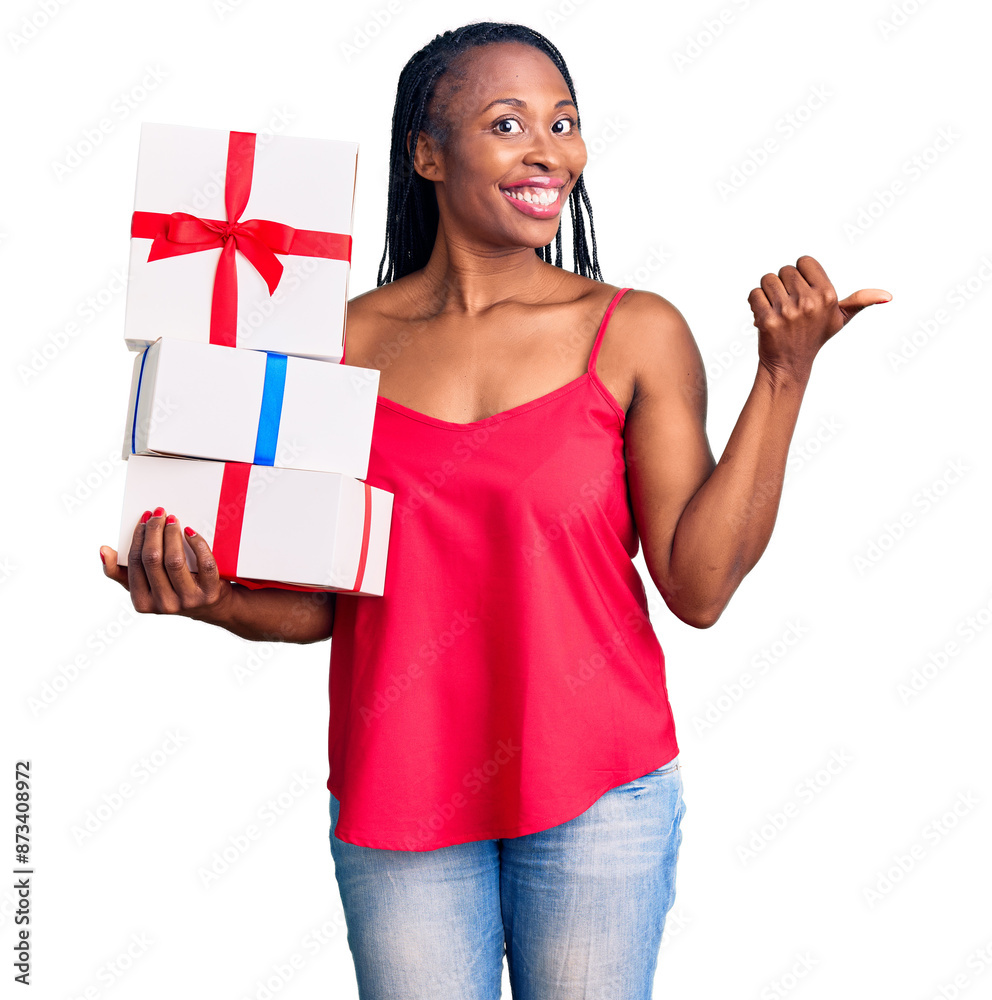 Sticker Young african american woman holding gift pointing thumb up to the side smiling happy with open mouth