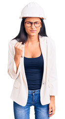 Young beautiful latin girl wearing architect hardhat and glasses angry and mad raising fist frustrated and furious while shouting with anger. rage and aggressive concept.