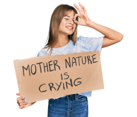 Teenager caucasian girl holding mother nature is crying protest cardboard banner smiling happy doing ok sign with hand on eye looking through fingers
