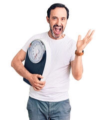 Middle age handsome man holding weighing machine celebrating victory with happy smile and winner expression with raised hands