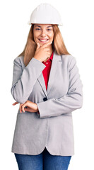 Beautiful young woman wearing architect hardhat looking confident at the camera smiling with crossed arms and hand raised on chin. thinking positive.