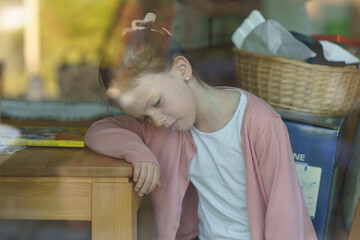 Photo of a little tired girl in a pink blouse bowing her head while sitting at a wooden table...