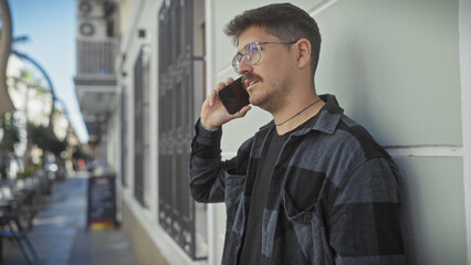 Handsome young hispanic man with glasses and a moustache talking on the phone on a sunny city street