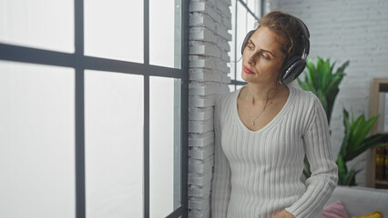 A thoughtful young woman enjoys music with headphones in a cozy modern living room with plants and natural light.