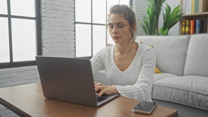 A focused young woman using a laptop in a modern living room with natural light and minimalistic decor.