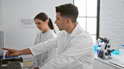 A man and woman in lab coats work together in a laboratory, examining data on a computer screen.