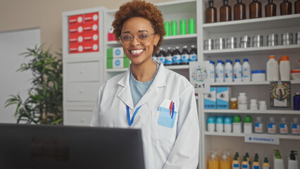 Smiling african american woman pharmacist wearing glasses stands in pharmacy with shelves of medications.