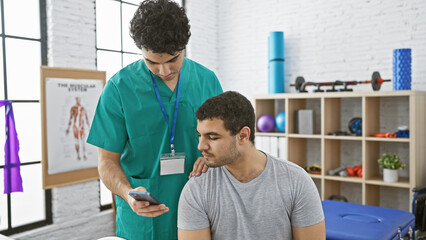 Fototapeta premium A male therapist in scrubs uses a smartphone in a modern physiotherapy clinic with a male patient.