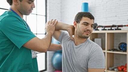 Therapist in scrubs performing shoulder physiotherapy on male patient inside a well-equipped clinic.