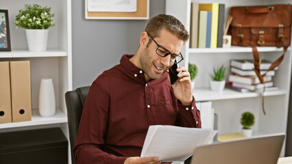 Fototapeta premium Smiling hispanic man in an office engaging in business over the phone while reviewing documents.