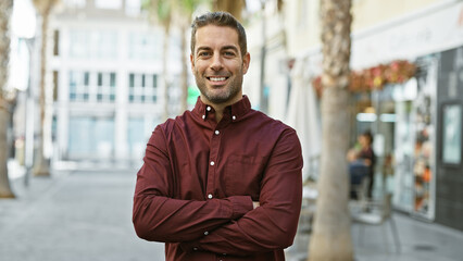 Obraz premium Smiling, confident hispanic man with a beard stands with his arms crossed on a sunny city street.