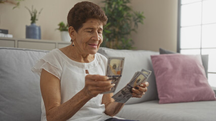 An elderly hispanic woman with short hair counts a stack of us dollar bills while sitting on a couch in a cozy living room.