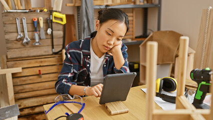 A focused asian woman using a tablet in a cluttered carpentry workshop surrounded by tools and wood.