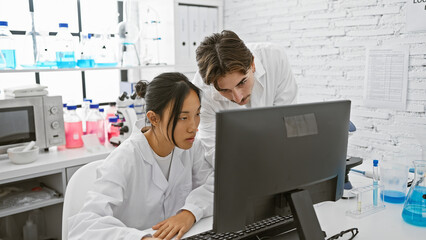 Man and woman scientists in laboratory analyzing data on computer amidst equipment.