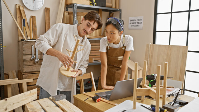 Two carpenters, a man and a woman, working together in a well-lit woodshop, discussing a project with a laptop on the workbench