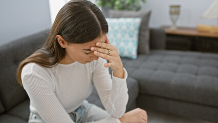 A stressed young woman indoors touches her forehead, showcasing worry or a headache in a comfortable living room.