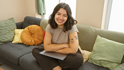 Obraz premium Smiling young hispanic woman sitting comfortably at home on a couch with colorful pillows and a laptop.