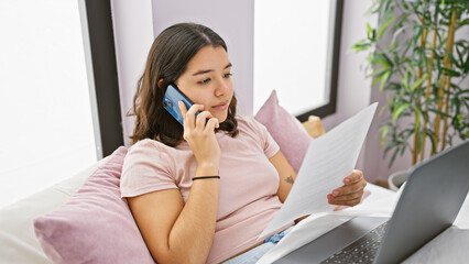 Hispanic woman multitasking with phone and document in a home bedroom setting, exuding a professional yet relaxed vibe.