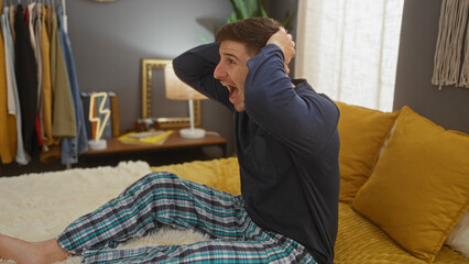 Fototapeta premium A surprised young hispanic man sits on a bed with his hands on his head, in a cozy bedroom with warm lighting, surrounded by decor and clothes in the background.