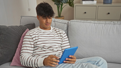 A handsome young hispanic man enjoys a tablet in his cozy living room, seated comfortably on a modern sofa, highlighting the relaxed indoor ambiance of his stylish home.