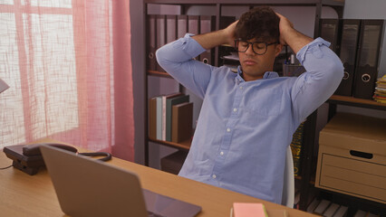 A young hispanic man in an office sits at a desk with his hands on his head, looking worried and stressed, surrounded by office supplies and shelves in an indoor workplace setting.