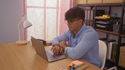 Handsome hispanic man working on a laptop in an office setting with organized shelves and desk accessories.