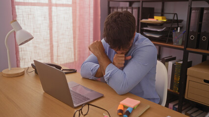 A young hispanic man coughs at his workplace desk in an indoor office setting, surrounded by a laptop, lamp, and office supplies.