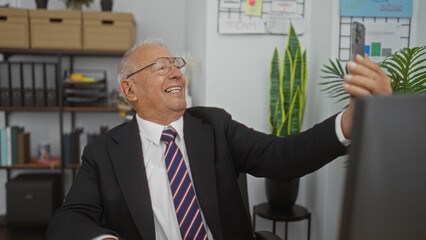 An elderly man with grey hair and glasses, taking a selfie in a modern office workspace with indoor plants and organized shelves in the background.