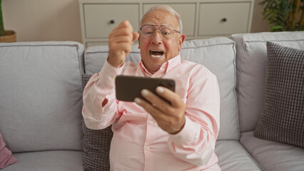 A senior caucasian man with grey hair is seated indoors in a living room, looking excited and holding a smartphone.