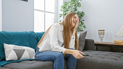 Obraz premium Laughing young blonde woman holding a remote in a cozy living room.