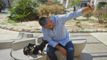 An adult hispanic man takes a selfie with two small chihuahuas in an outdoor urban park.